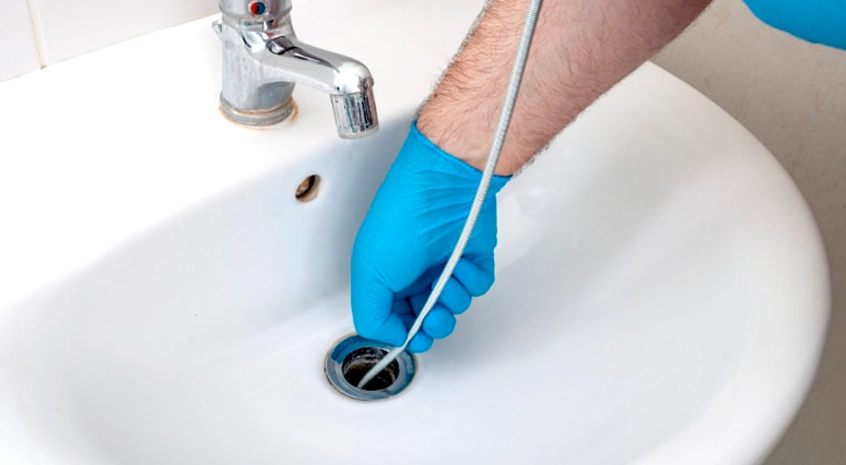 Hand wearing blue glove clearing bathroom sink drain with cable tool.